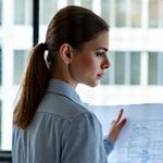 Nagy Éva, Vezető Építész, egy tervrajz felett elmélkedik, koncentrált arccal, modern építészirodában.